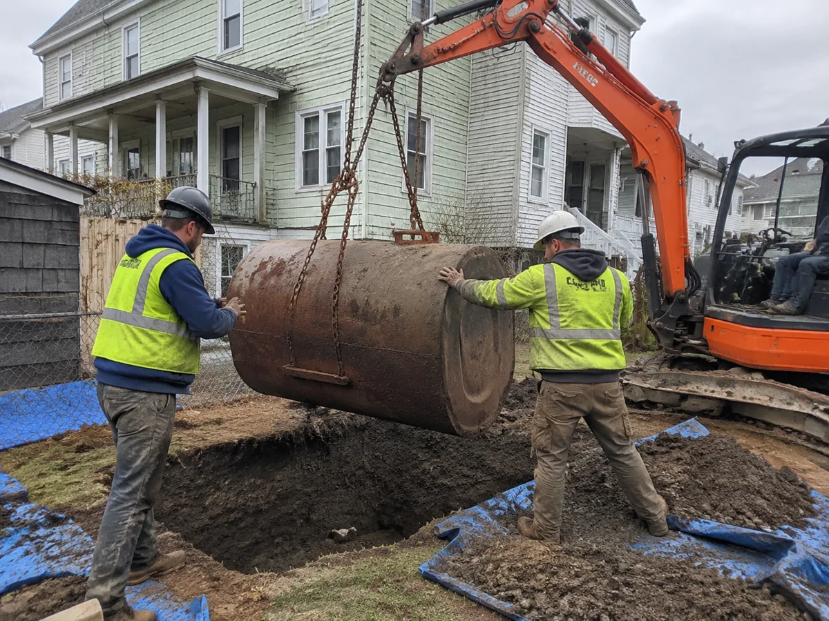 Oil tank removal New Bedford MA crew lifting a 275-gallon steel tank out of a backyard with a mini excavator